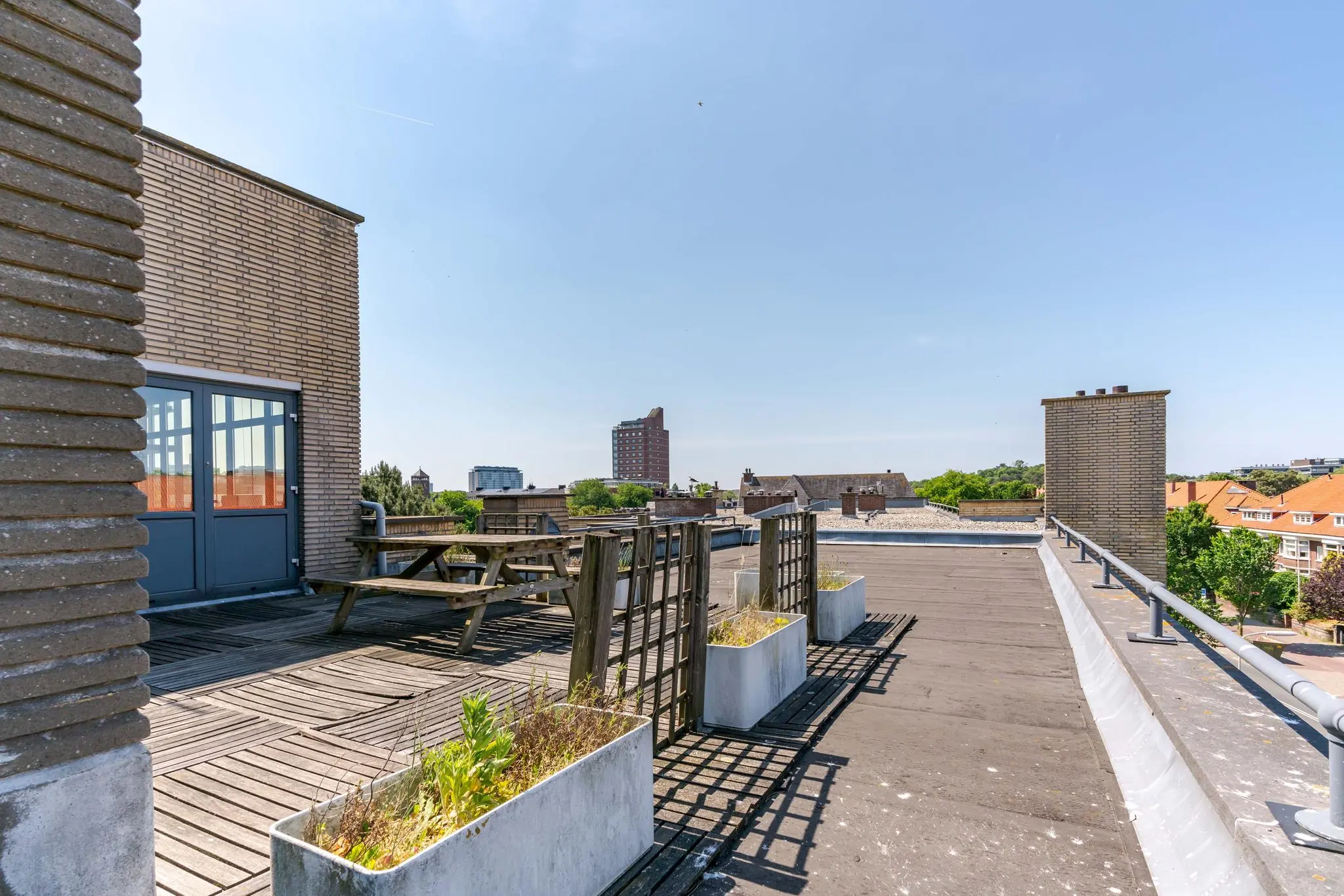 Dakterras aan de Van Alkemadelaan met houten vlonder, picknicktafel, plantenbakken en uitzicht op omliggende gebouwen.